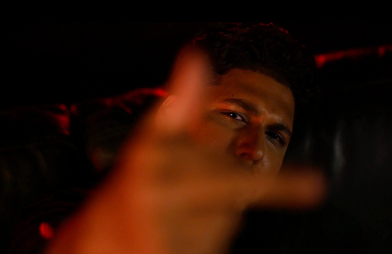 ISSA G lit in red, looking into the camera past a blurred hand reaching toward the lens